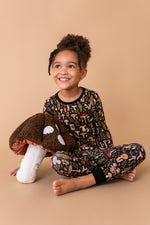A young girl smiles cross-legged on a beige background wearing Jammie Session’s Mystic Mushrooms Long Sleeve PJ's—soft, hypoallergenic kids pajamas with a whimsical plants and mushrooms print, perfect for sensitive skin. She hugs a plush mushroom toy.