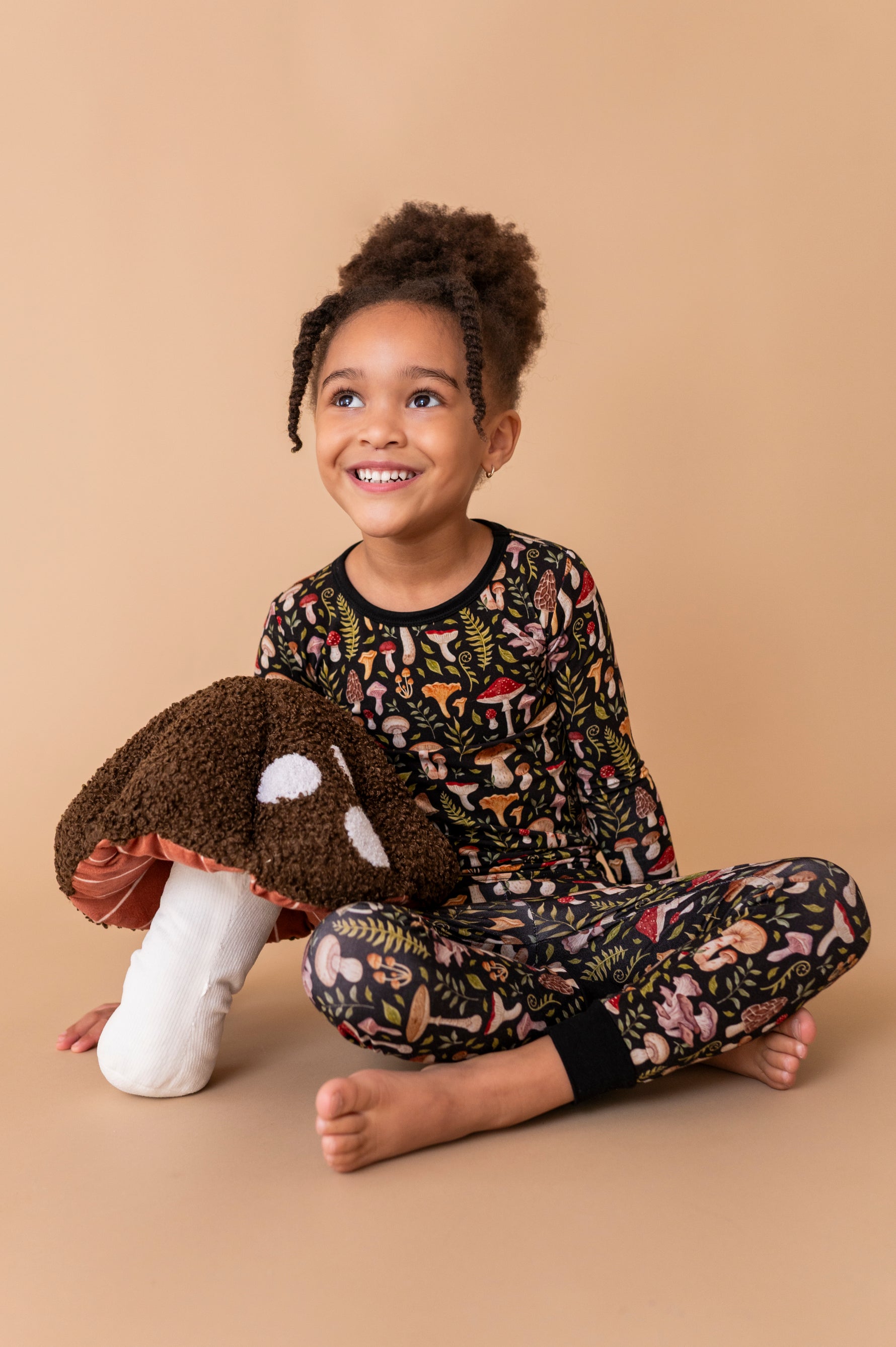 A young girl smiles cross-legged on a beige background wearing Jammie Session’s Mystic Mushrooms Long Sleeve PJ's—soft, hypoallergenic kids pajamas with a whimsical plants and mushrooms print, perfect for sensitive skin. She hugs a plush mushroom toy.