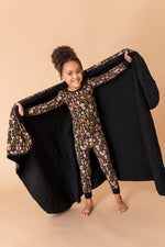 A young girl smiles barefoot on a beige background, holding open the Jammie Session Mystic Mushrooms Toddler Quilted Blanket behind her while wearing patterned pajamas.
