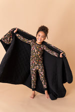 A young child in patterned pajamas stands barefoot on a tan background, smiling at the camera while holding open a Jammie Session Mystic Mushrooms Toddler Quilted Blanket behind them.