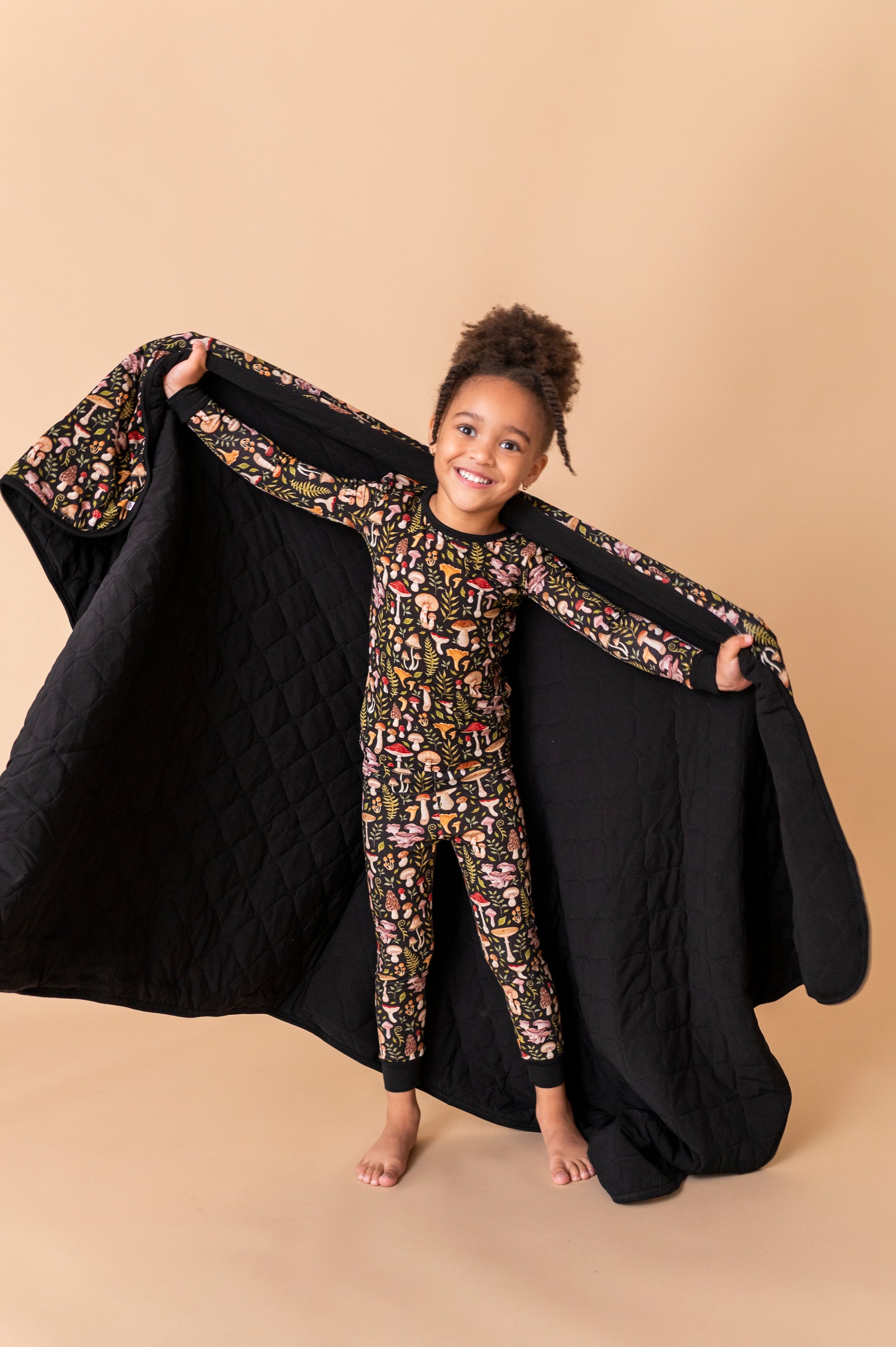 A young child in patterned pajamas stands barefoot on a tan background, smiling at the camera while holding open a Jammie Session Mystic Mushrooms Toddler Quilted Blanket behind them.