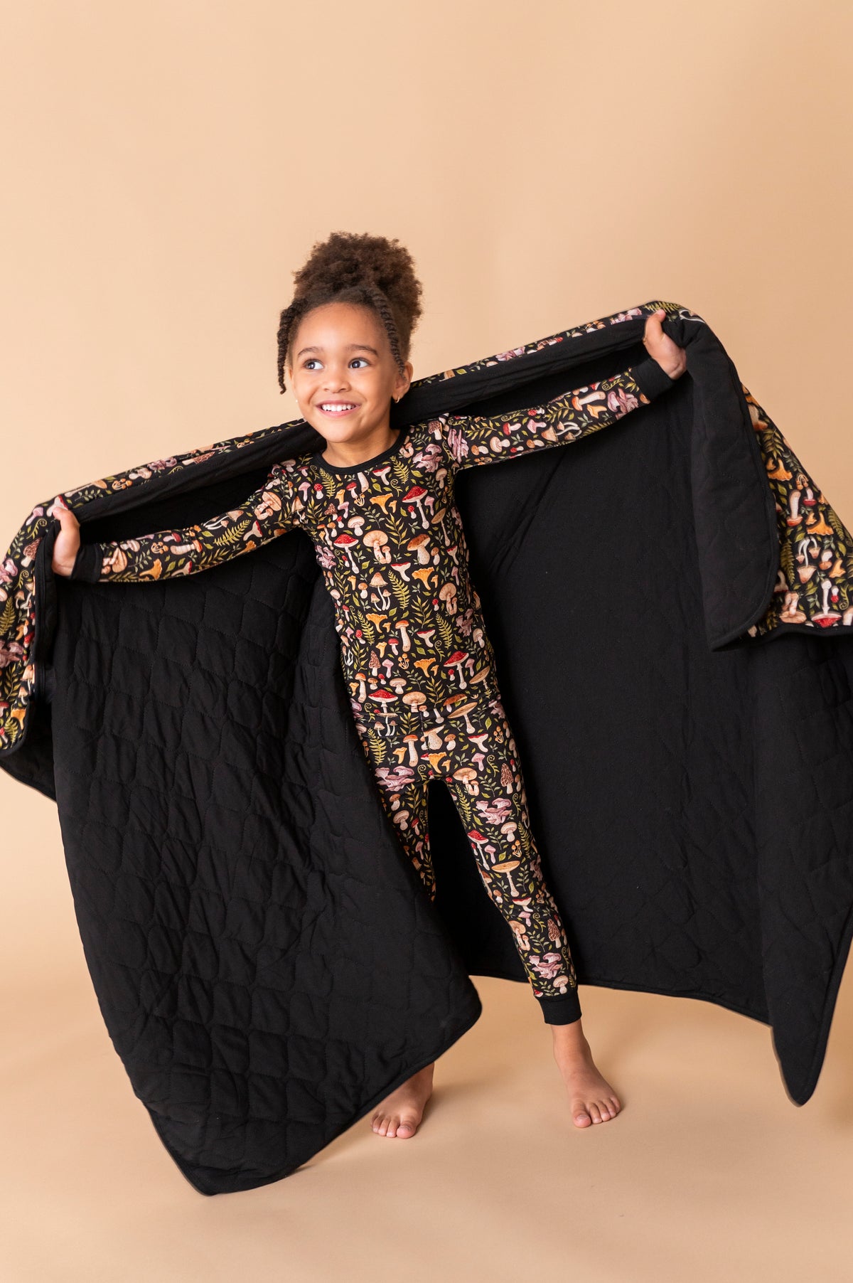 A young child in patterned pajamas stands barefoot, smiling and holding open a Jammie Session Mystic Mushrooms Toddler Quilted Blanket against a beige background.