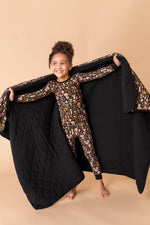 A young child in patterned pajamas stands barefoot, smiling and holding open a Jammie Session Mystic Mushrooms Toddler Quilted Blanket against a beige background.