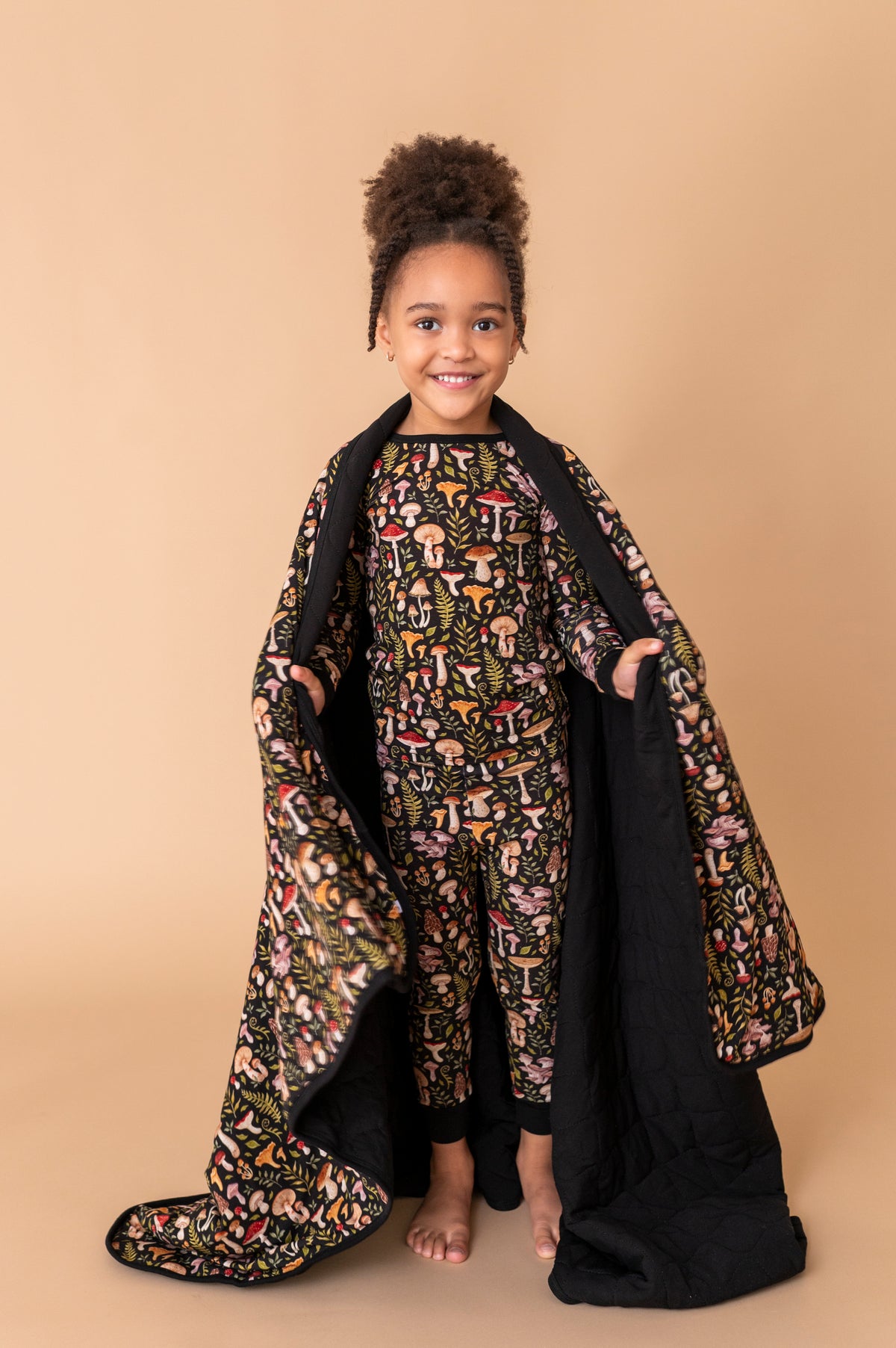 A young child smiles at the camera, standing barefoot on a beige backdrop while holding the Jammie Session Mystic Mushrooms Toddler Quilted Blanket and wearing matching patterned pajamas.