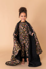 A young child smiles at the camera, standing barefoot on a beige backdrop while holding the Jammie Session Mystic Mushrooms Toddler Quilted Blanket and wearing matching patterned pajamas.