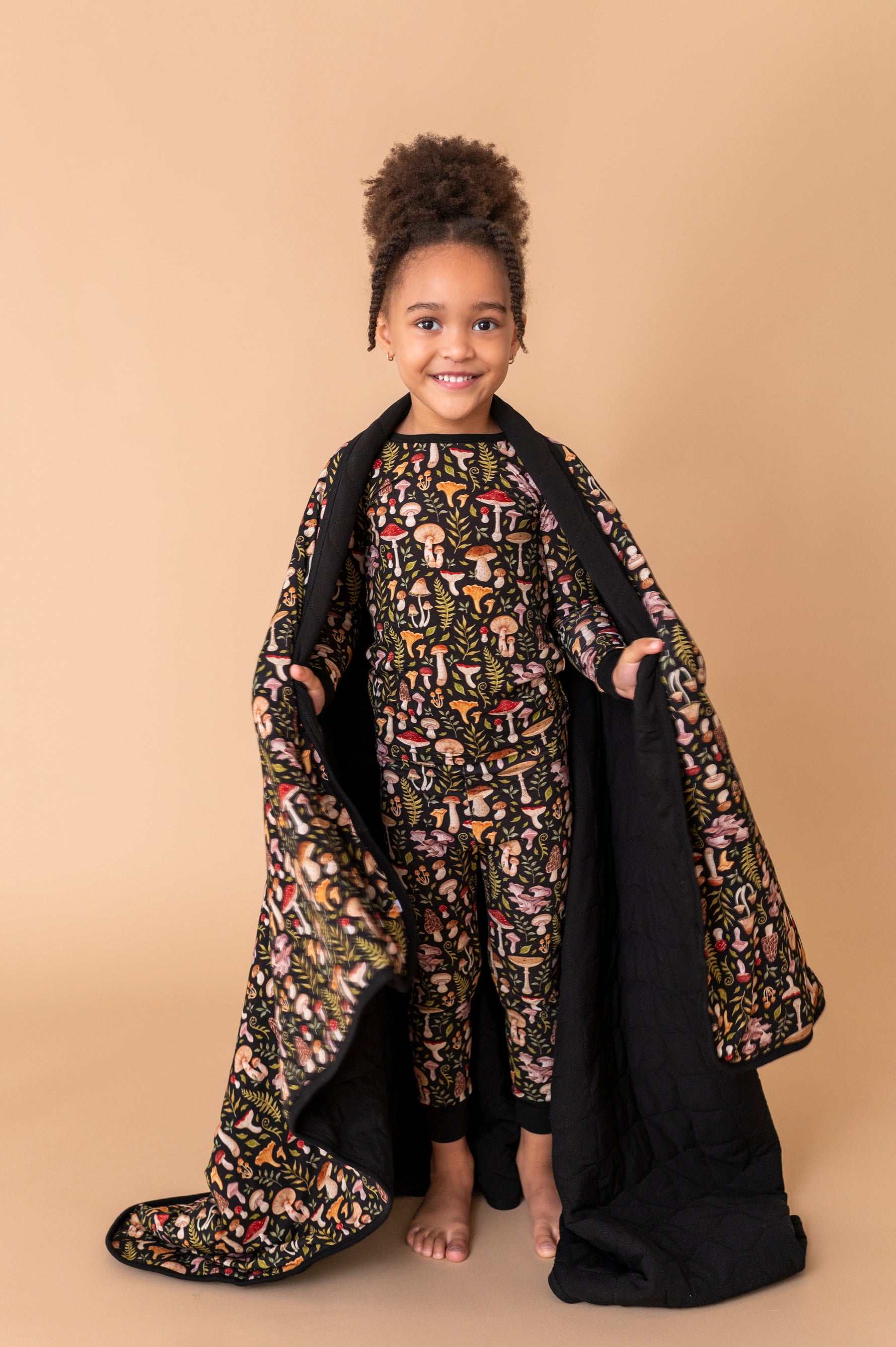 A young child smiles at the camera, standing barefoot on a beige backdrop while holding the Jammie Session Mystic Mushrooms Toddler Quilted Blanket and wearing matching patterned pajamas.
