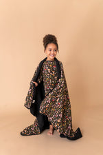 A young child smiles barefoot, wearing a patterned dress and holding the Jammie Session Mystic Mushrooms Toddler Quilted Blanket against a plain beige background.