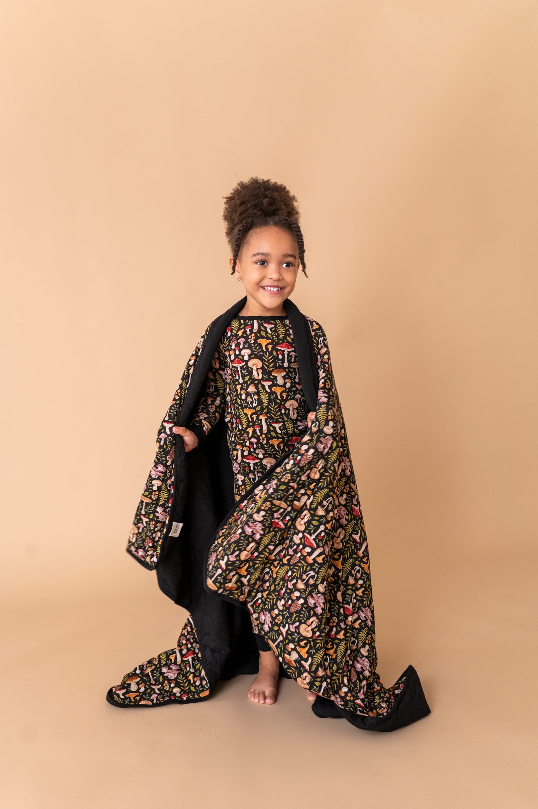 A young child smiles barefoot, wearing a patterned dress and holding the Jammie Session Mystic Mushrooms Toddler Quilted Blanket against a plain beige background.
