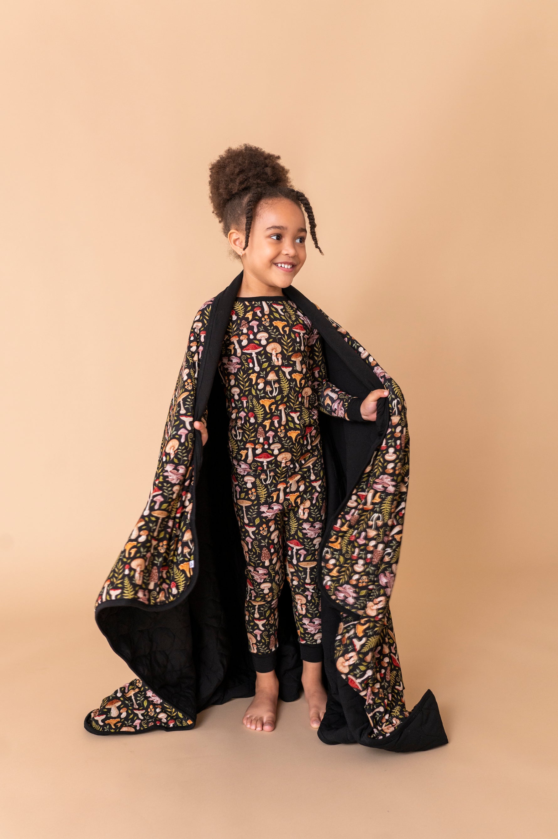 A barefoot child stands on a beige background, dressed in matching pajamas and draped in the Jammie Session Mystic Mushrooms Toddler Quilted Blanket featuring a coordinated pattern.