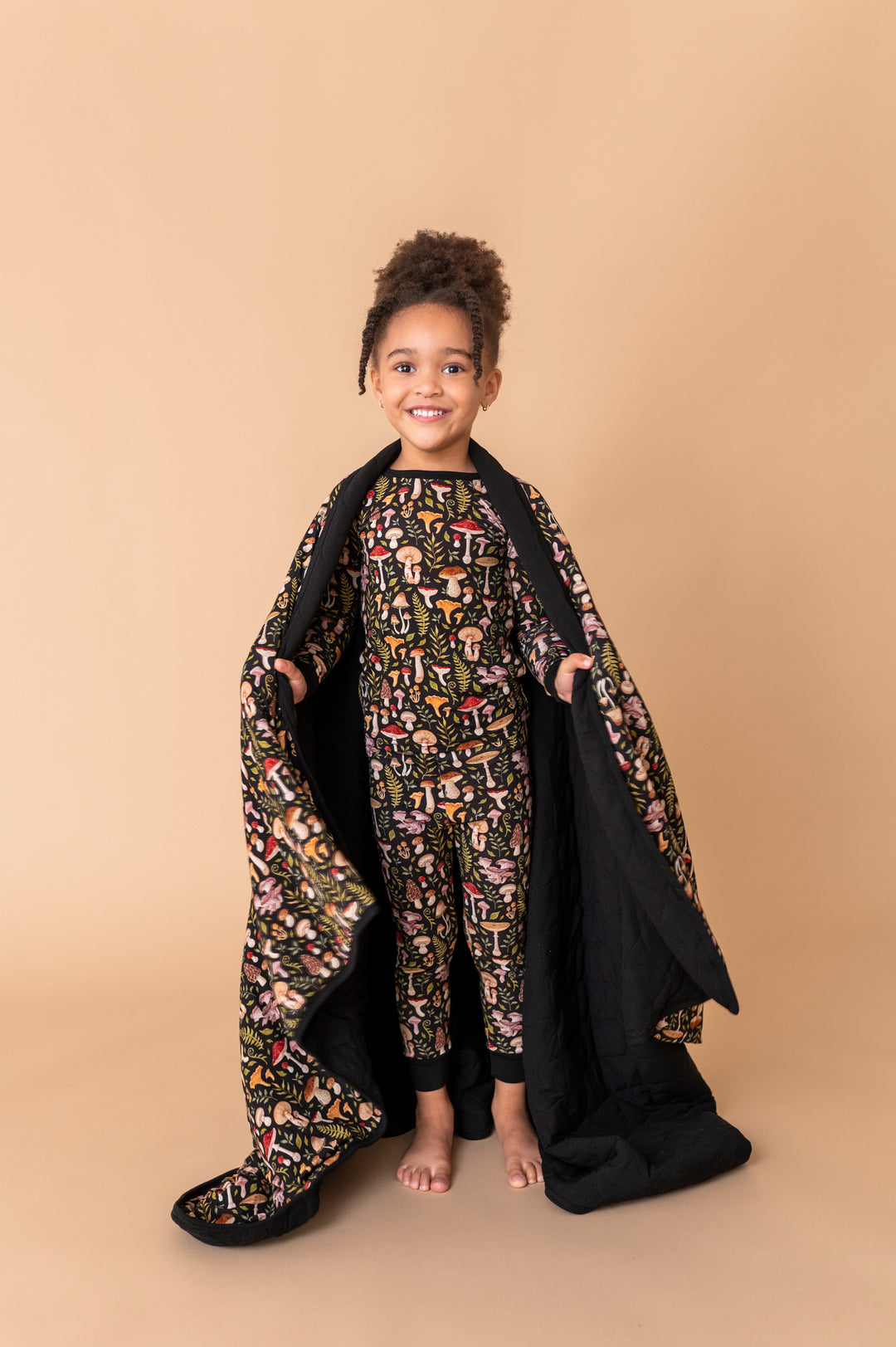 A young child smiles at the camera, standing barefoot on a beige background in matching pajamas while holding open the Jammie Session Mystic Mushrooms Toddler Quilted Blanket.
