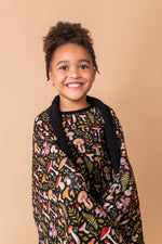 A young child smiles at the camera, wrapped in the Jammie Session Mystic Mushrooms Toddler Quilted Blanket, a black hypoallergenic blanket with a colorful mushroom and plant pattern, set against a beige background.