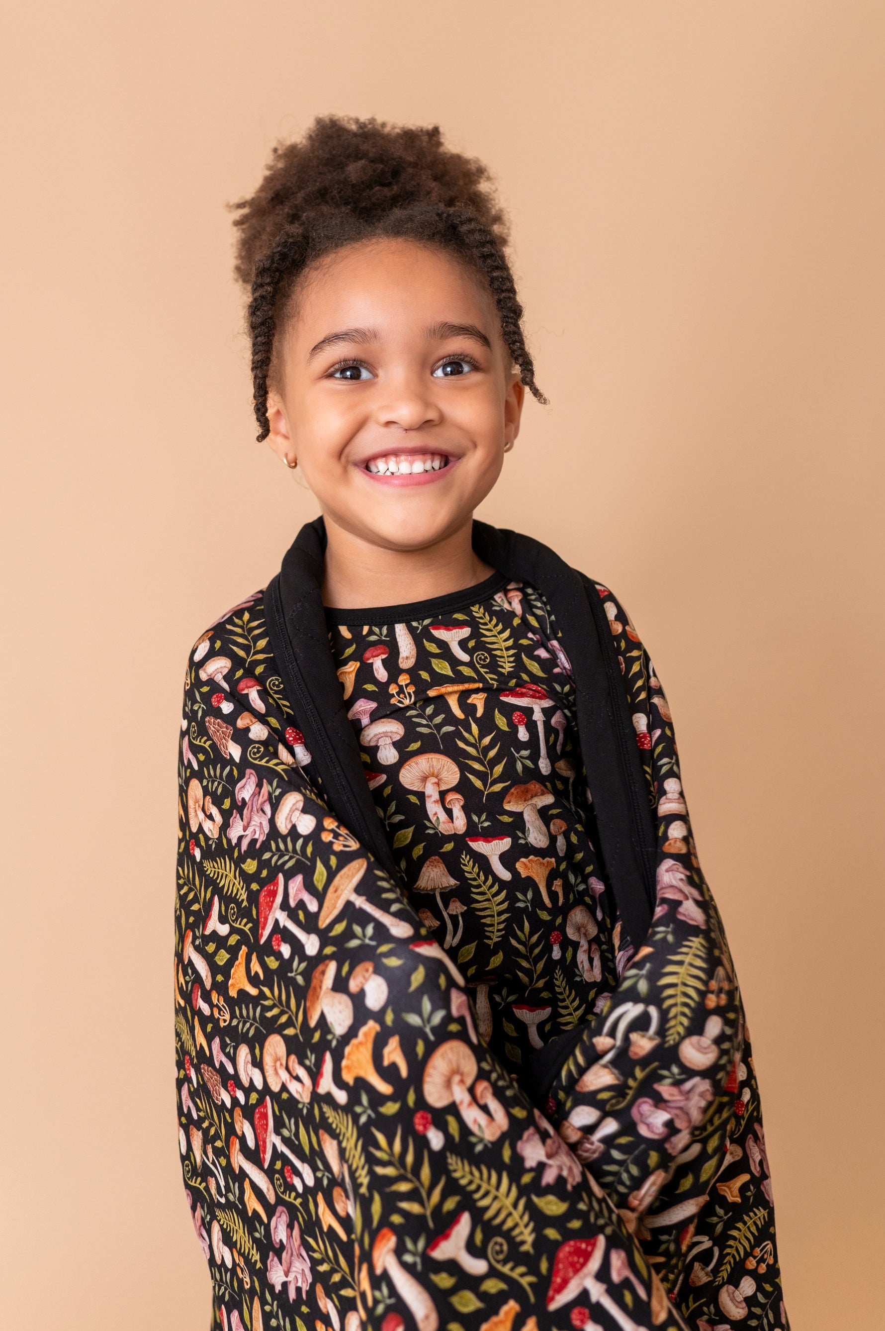 A young child smiles at the camera, wrapped in the Jammie Session Mystic Mushrooms Toddler Quilted Blanket, a black hypoallergenic blanket with a colorful mushroom and plant pattern, set against a beige background.