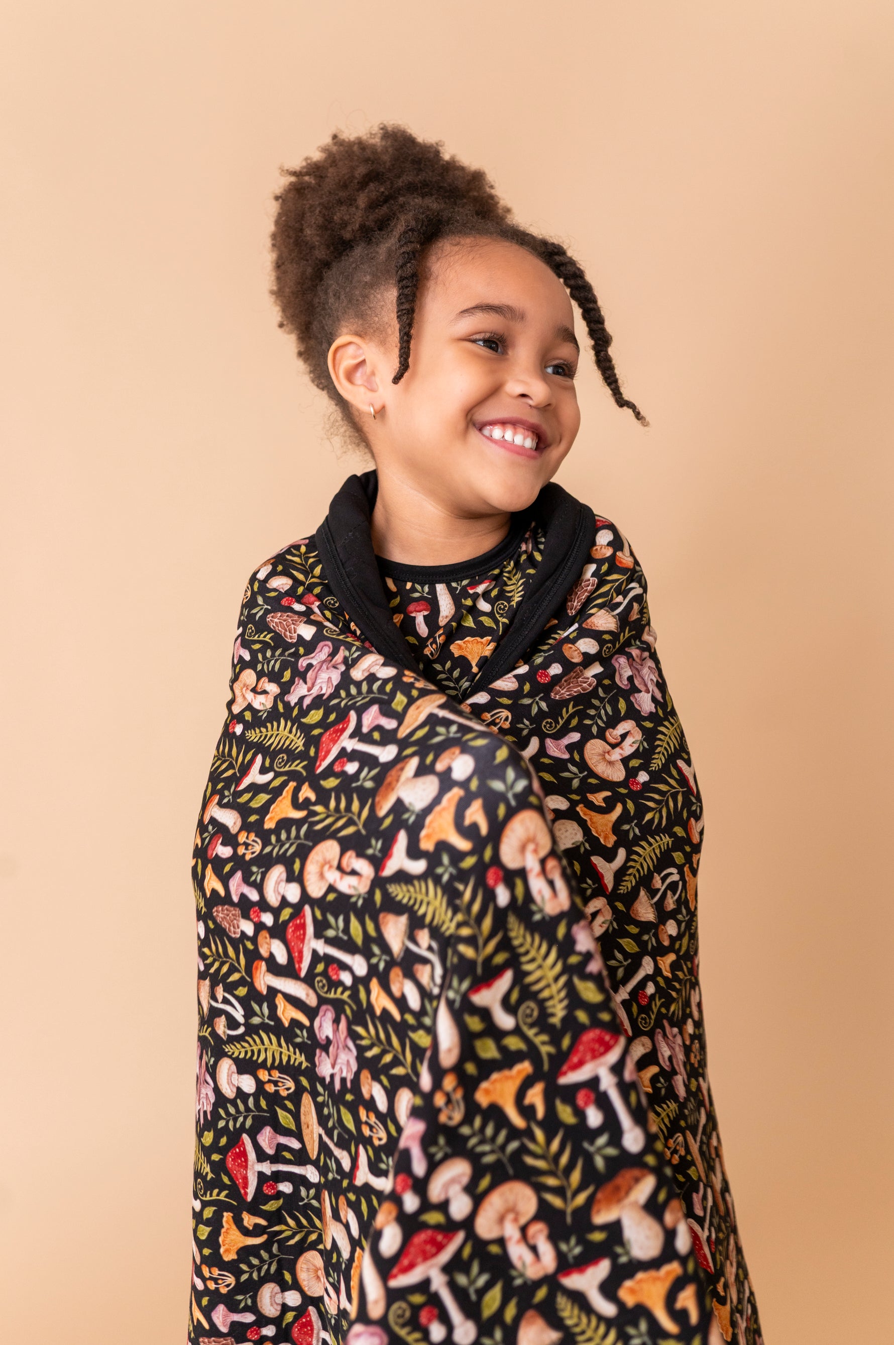 A smiling child with curly hair stands against a beige background, wrapped in the Jammie Session Mystic Mushrooms Toddler Quilted Blanket featuring a colorful mushroom and plant pattern.