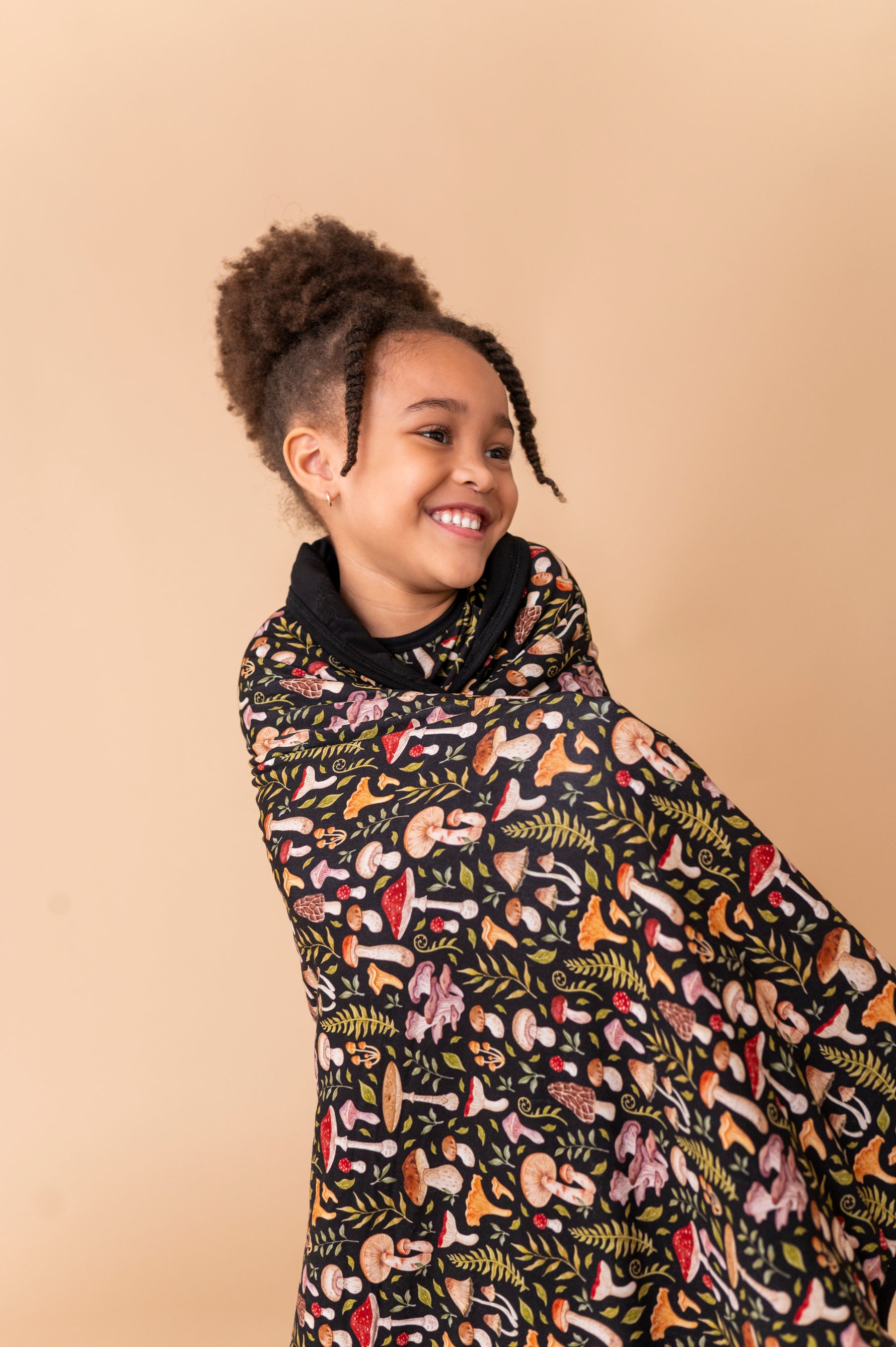 A young girl smiles, wrapped in the Jammie Session Mystic Mushrooms Toddler Quilted Blanket with colorful botanical and mushroom patterns, standing before a plain beige background.
