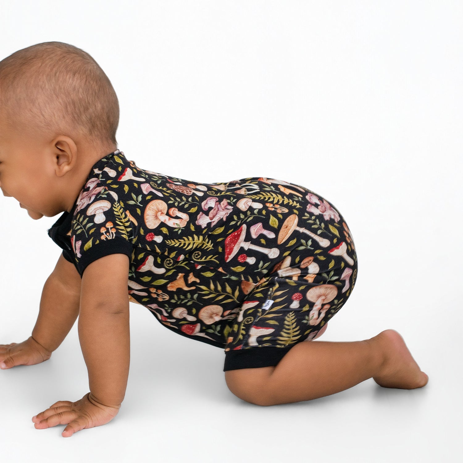 A baby wearing the Jammie Session Mystic Mushrooms Short Sleeve Romper, featuring a vibrant mushroom and foliage print, crawls on a plain white background.