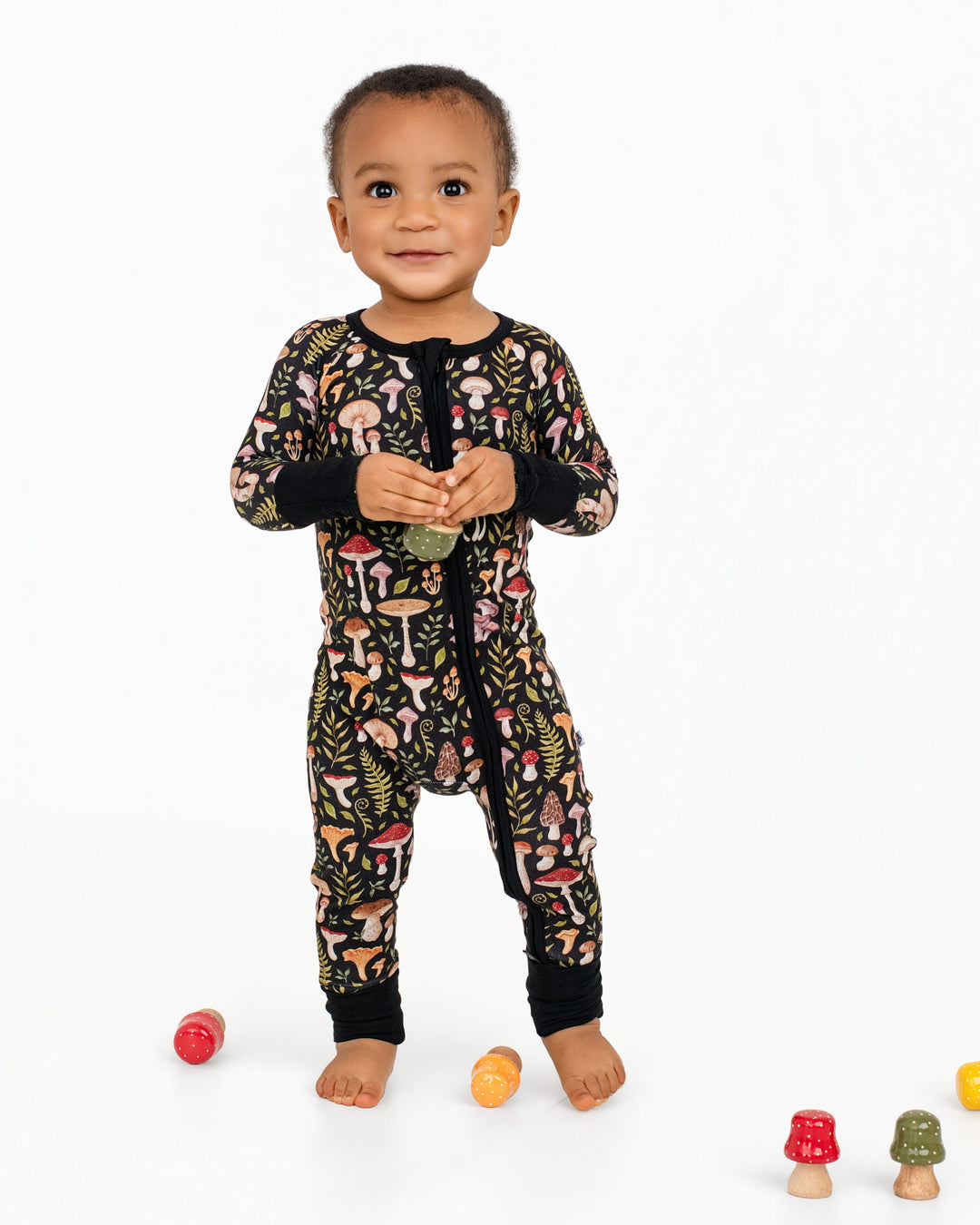 A toddler wears the black Mystic Mushrooms Romper by Jammie Session, featuring a colorful mushroom pattern, and stands on a white background holding a small object with toy mushrooms scattered nearby.