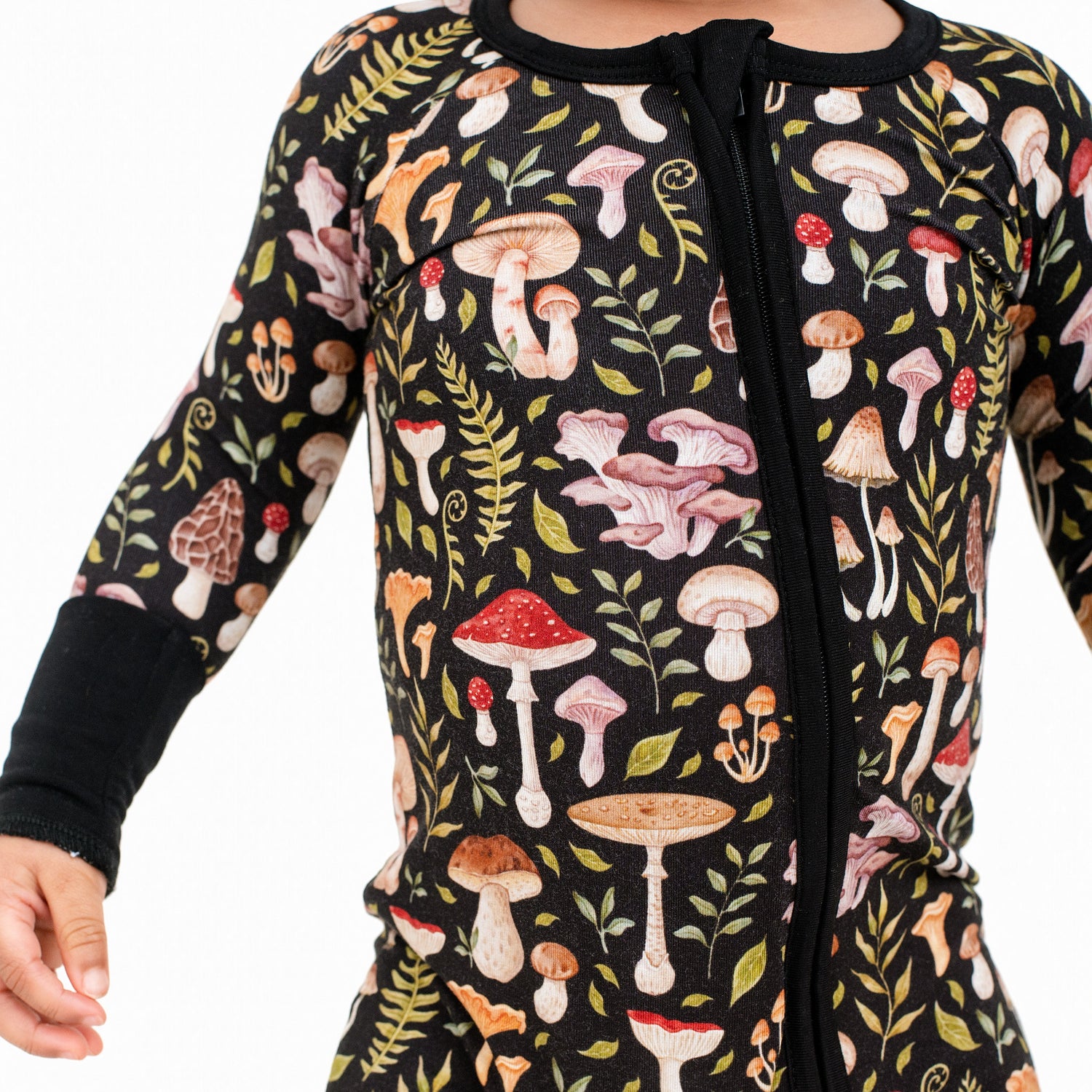 A child wearing the Jammie Session Mystic Mushrooms Romper, a black long sleeve outfit decorated with a vibrant mushroom and foliage design.