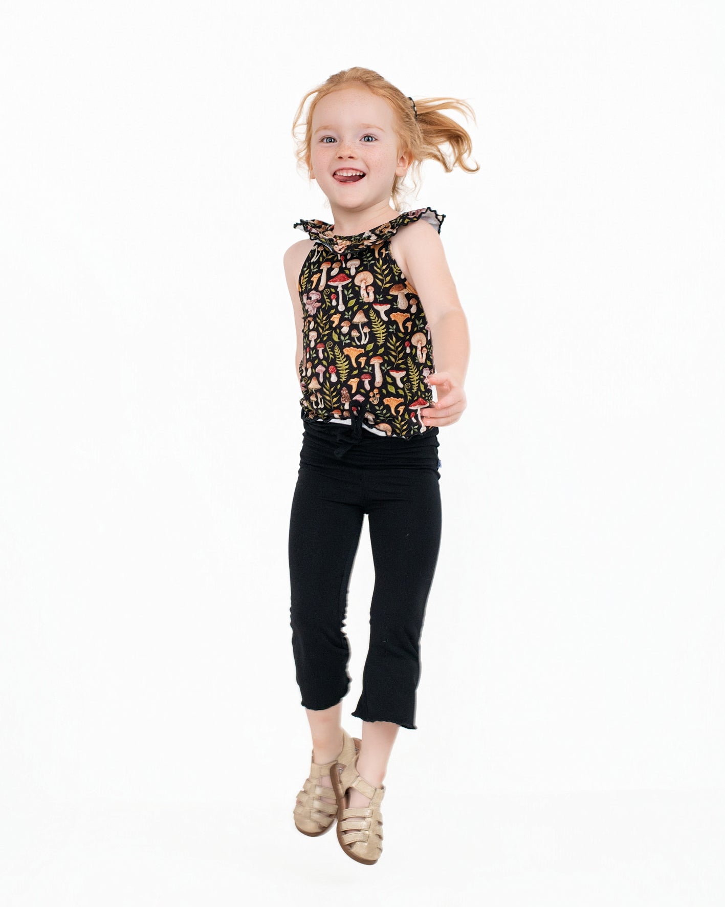A young girl with red hair wears the Jammie Session Mystic Mushrooms Bloomy Crop Set, captured mid-jump against a plain white background.