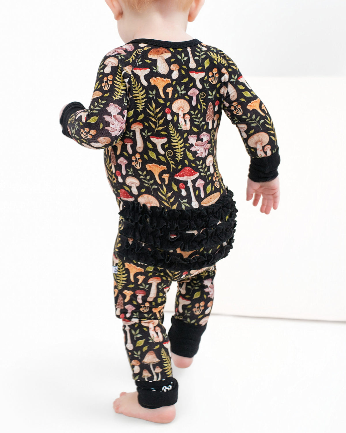 A toddler in Jammie Session's Mystic Mushrooms Ruffle Romper, a black bamboo outfit with a vibrant mushroom and fern pattern plus ruffled back, walks barefoot on a white surface.