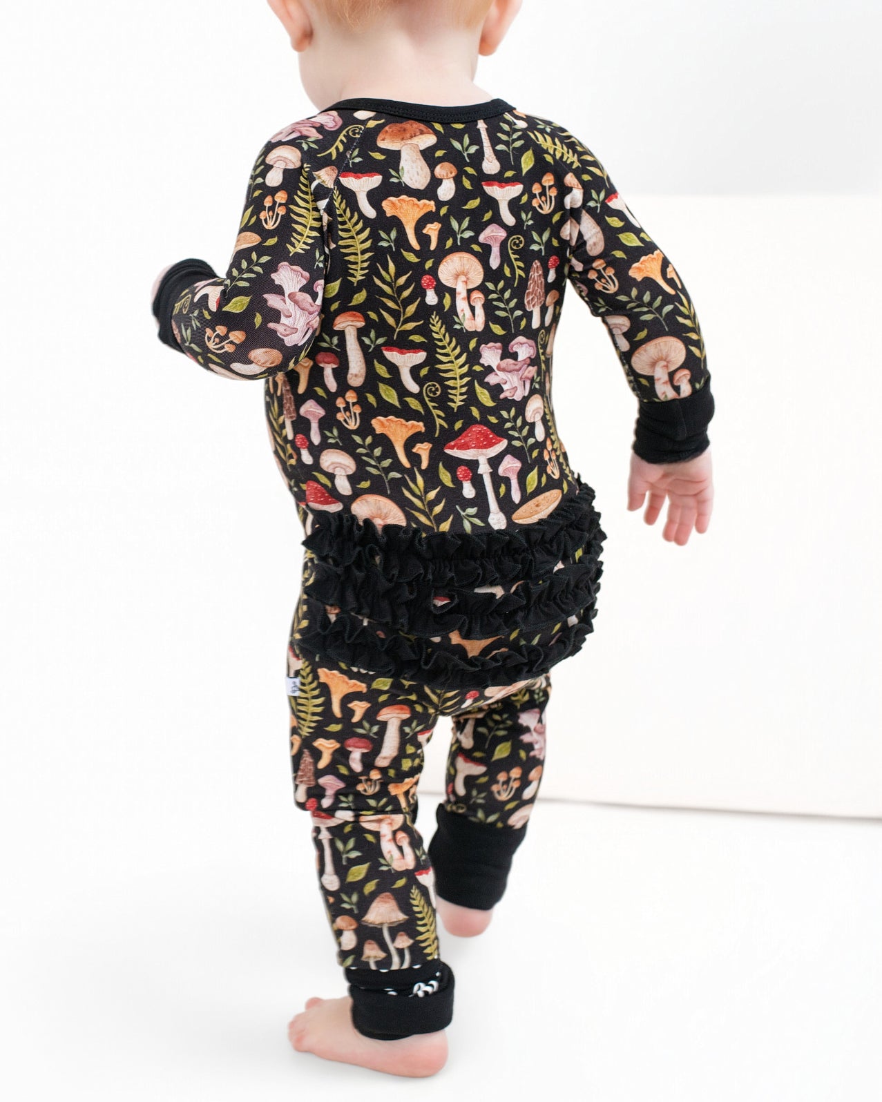 A toddler in Jammie Session's Mystic Mushrooms Ruffle Romper, a black bamboo outfit with a vibrant mushroom and fern pattern plus ruffled back, walks barefoot on a white surface.
