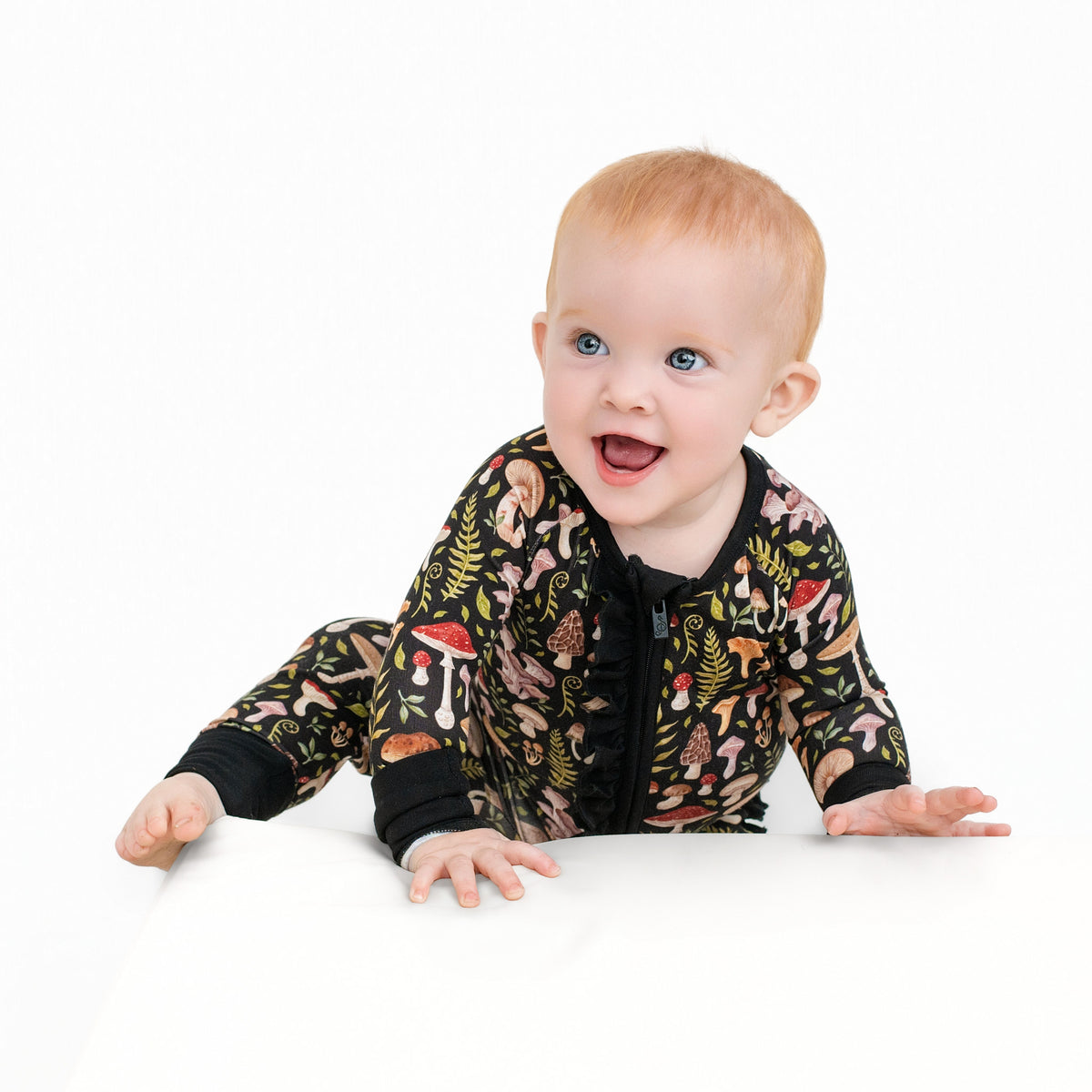A baby with light hair crawls on a white surface, dressed in the Jammie Session Mystic Mushrooms Ruffle Romper—a black long sleeve romper featuring colorful mushroom and plant prints—against a plain white background.