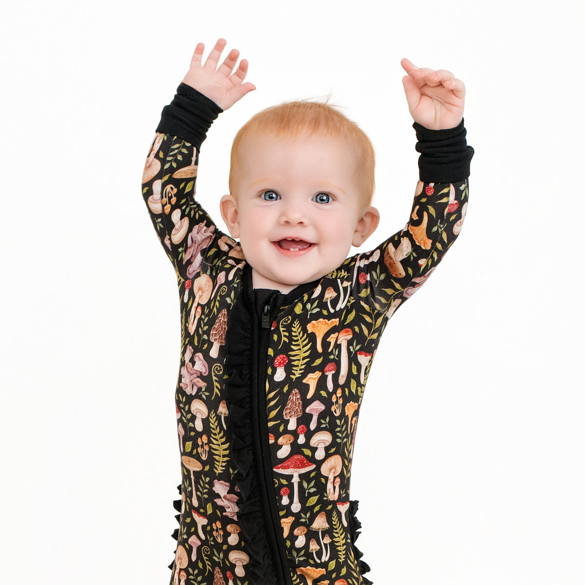 A light-haired baby smiles and raises both arms while wearing the Jammie Session Mystic Mushrooms Ruffle Romper, a black long sleeve outfit decorated with colorful mushrooms and leaves, against a white background.