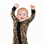 A light-haired baby smiles and raises both arms while wearing the Jammie Session Mystic Mushrooms Ruffle Romper, a black long sleeve outfit decorated with colorful mushrooms and leaves, against a white background.