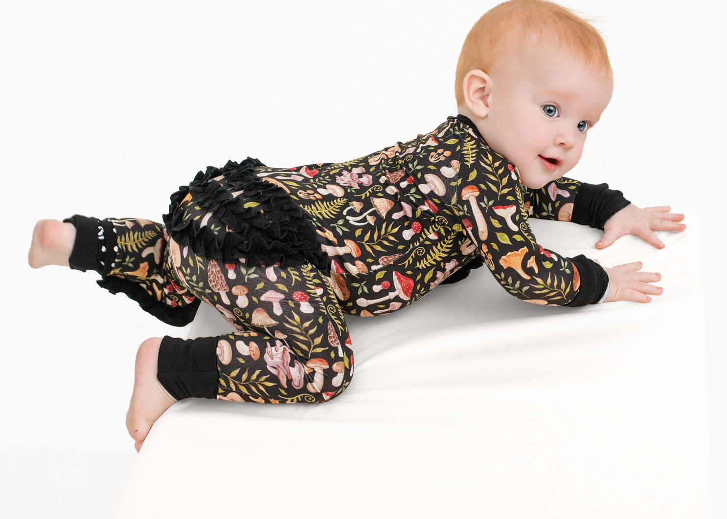 A light-skinned, red-haired baby crawls on a white surface wearing the Jammie Session Mystic Mushrooms Ruffle Romper, a dark patterned long sleeve outfit with ruffles on the back.