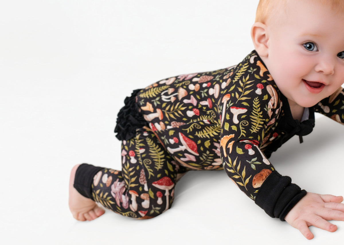 A baby crawls on a white surface in the adorable Mystic Mushrooms Ruffle Romper by Jammie Session, featuring a long-sleeve design with a colorful mushroom and foliage pattern.
