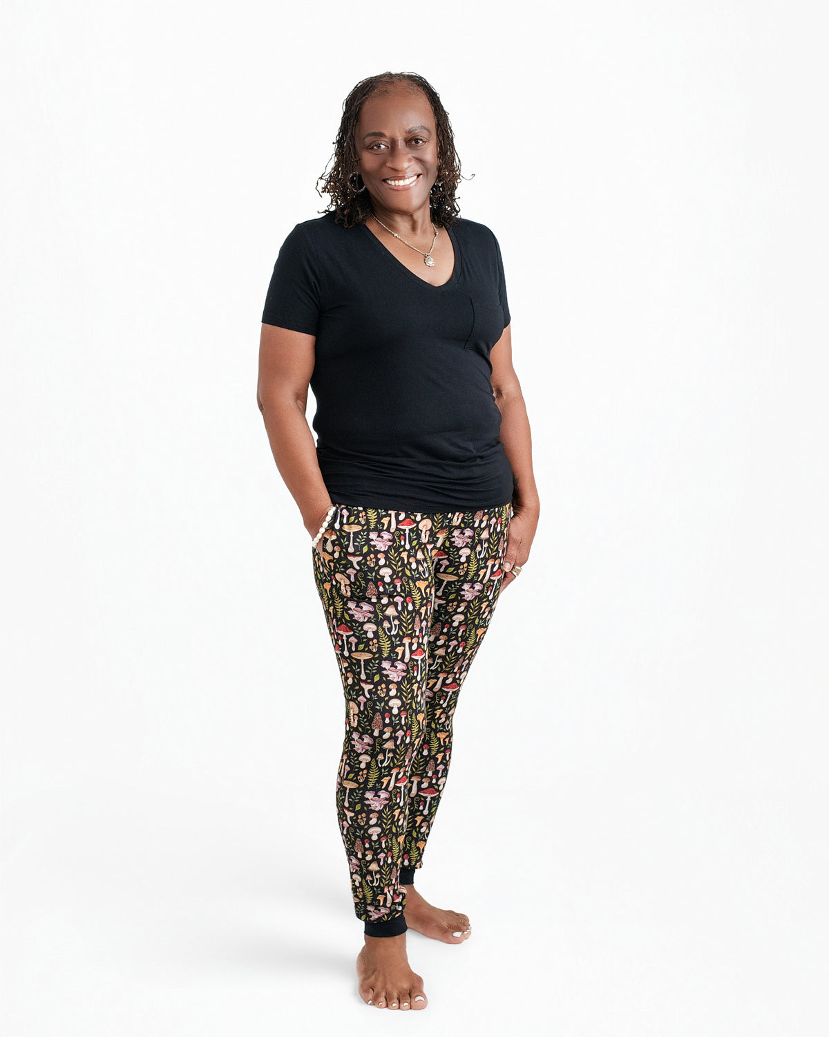 A woman stands barefoot against a white backdrop, smiling at the camera with her hands in her pockets, wearing Jammie Session's Mystic Mushrooms Women's Jogger Pants and a black t-shirt.
