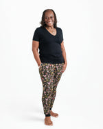 A woman stands barefoot against a white backdrop, smiling at the camera with her hands in her pockets, wearing Jammie Session's Mystic Mushrooms Women's Jogger Pants and a black t-shirt.