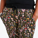 A person wearing Jammie Session's Mystic Mushrooms Women's Jogger Pants, featuring black fabric with vibrant mushroom and leaf patterns, stands with hands in pockets.