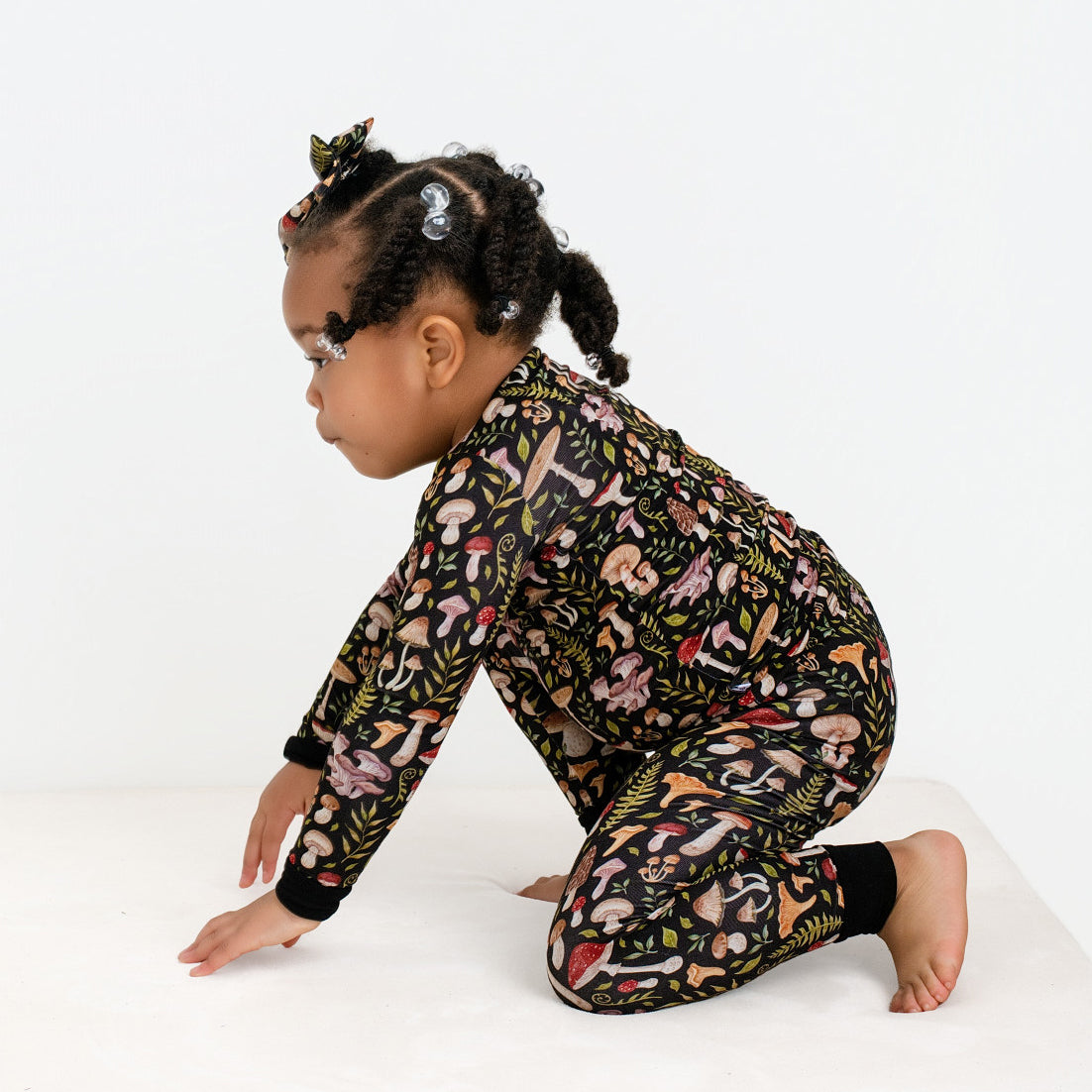 A young child in Jammie Session’s Mystic Mushrooms Long Sleeve PJ’s, featuring a buttery soft black floral pattern, kneels sideways on a white surface.