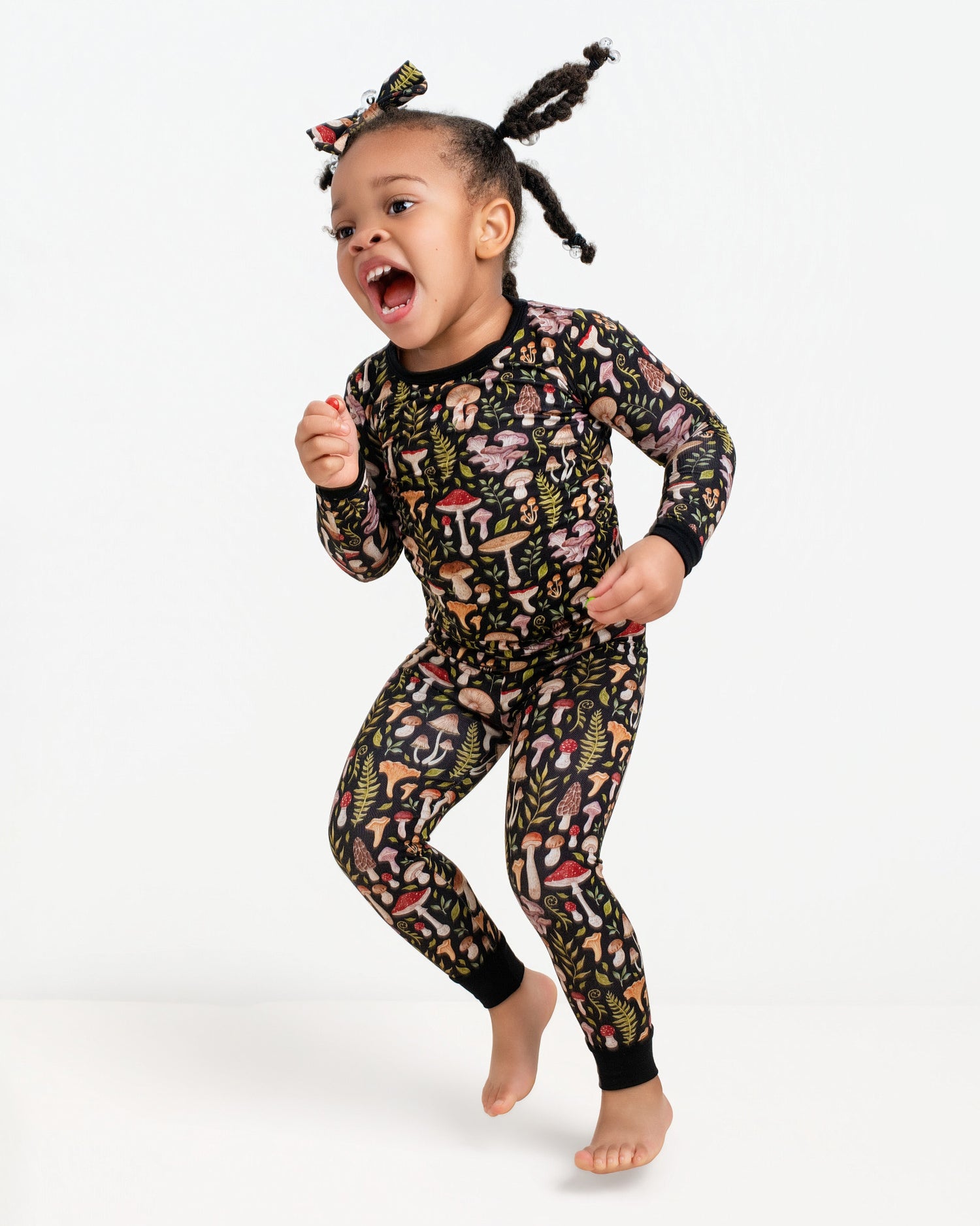A young child in Jammie Session’s Mystic Mushrooms Long Sleeve PJ's and matching headband jumps barefoot against a plain white background.