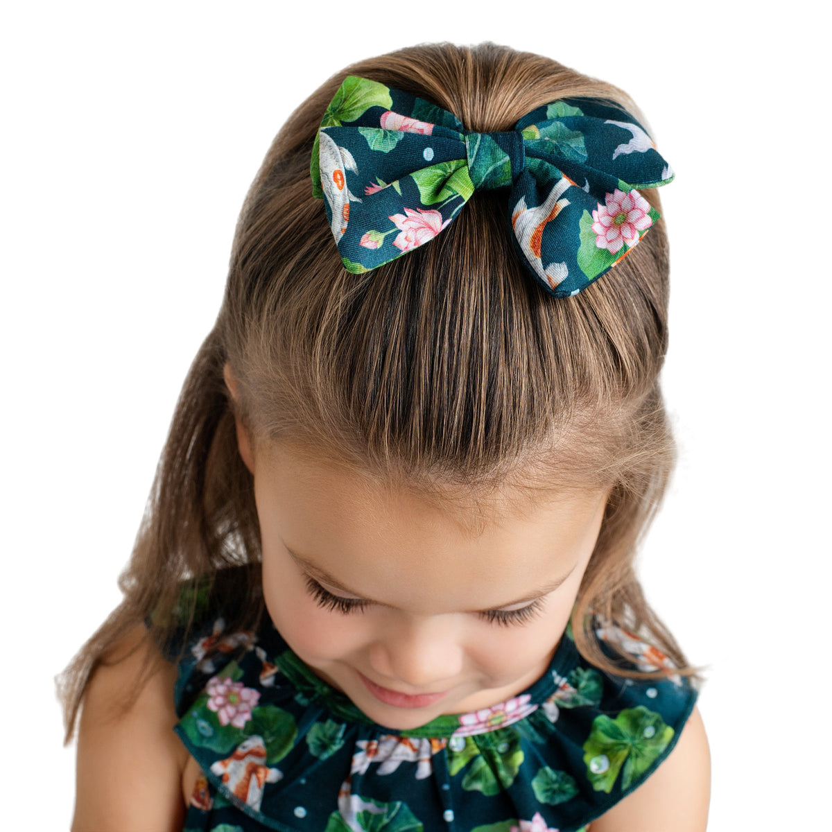 A young girl with light brown hair looks down, wearing the Jammie Session Bundle of Koi Bow featuring a floral and fish pattern that matches her dress, set against a white background.