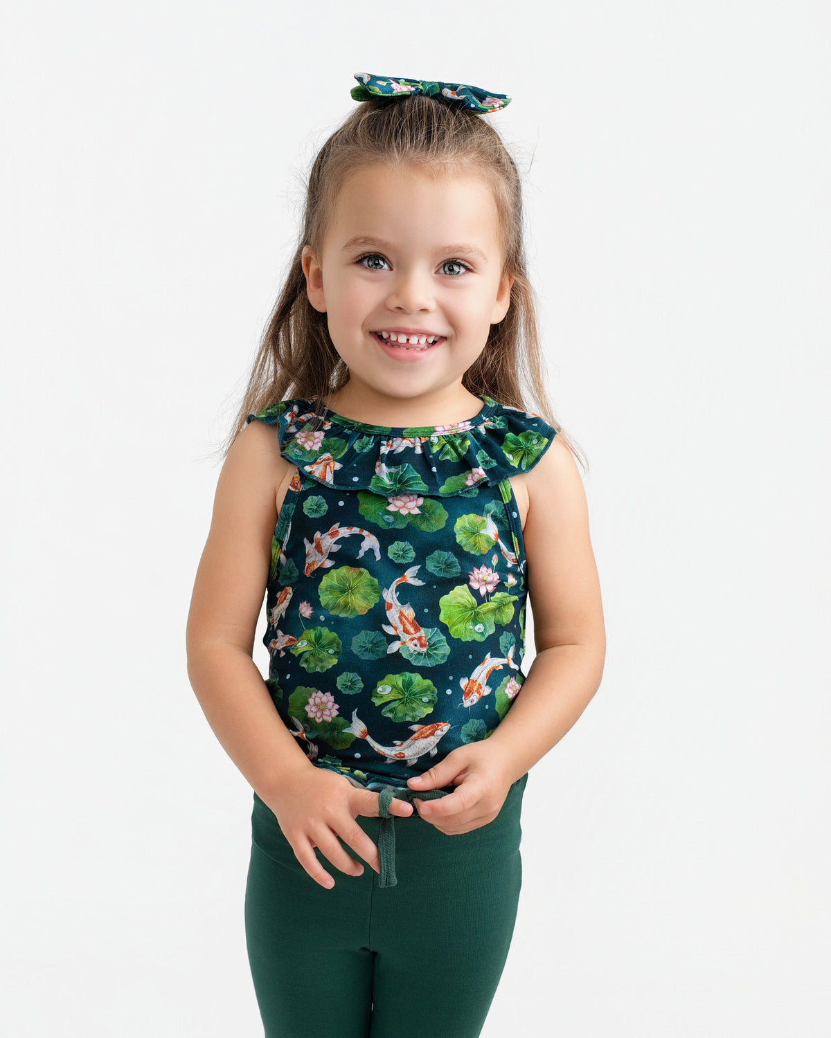 A young child with long brown hair wears the Jammie Session Bundle of Koi Bloomy Crop Set—featuring a matching floral top, hair bow, and dark green pants—standing and smiling against a plain white background.