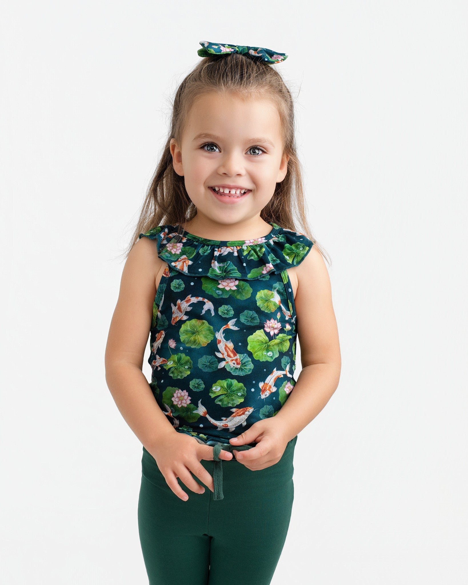 A young child with long brown hair wears the Jammie Session Bundle of Koi Bloomy Crop Set—featuring a matching floral top, hair bow, and dark green pants—standing and smiling against a plain white background.
