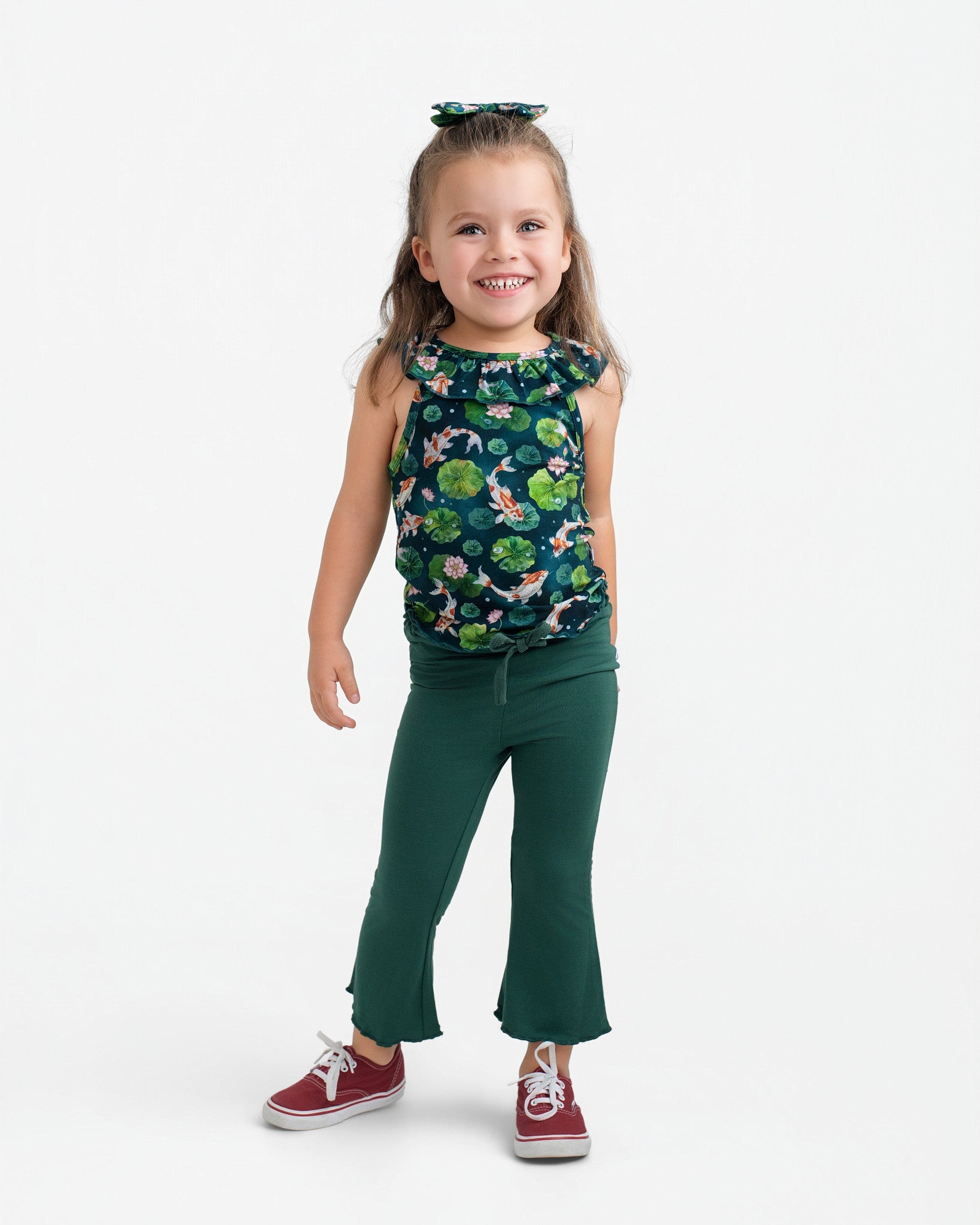 A young girl smiles in the Jammie Session Bundle of Koi Bloomy Crop Set—featuring a sleeveless floral top and green flared pants—paired with red sneakers and a coordinating hair bow against a plain white background.