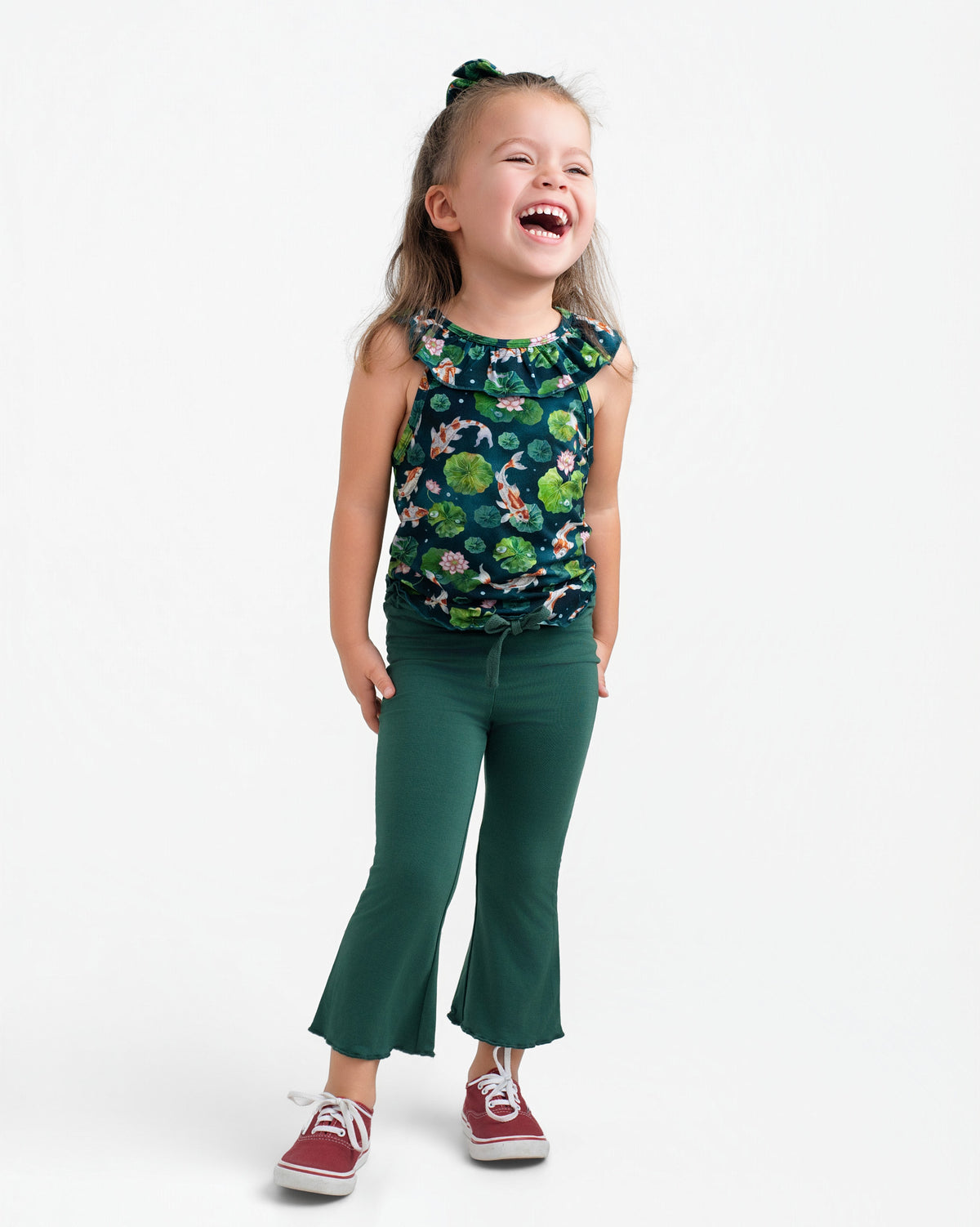 A young child smiles in the Jammie Session Bundle of Koi Bloomy Crop Set, featuring a green floral sleeveless top and matching pants, paired with red sneakers and a green hair tie, against a plain white background.