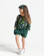 A young girl wears the Jammie Session Bundle of Koi Comfy Cloud Short Set—green-patterned top, matching shorts, and headband—with white shoes, standing with hands in pockets against a plain white background.