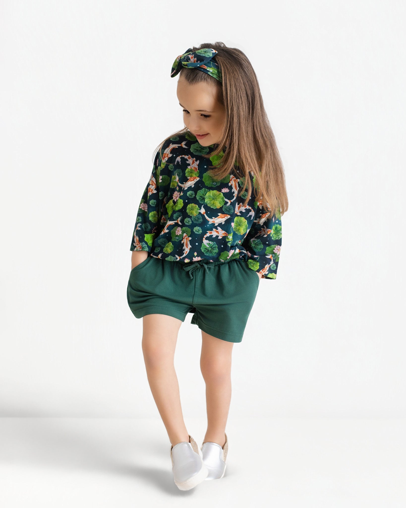 A young girl wears the Jammie Session Bundle of Koi Comfy Cloud Short Set—green-patterned top, matching shorts, and headband—with white shoes, standing with hands in pockets against a plain white background.