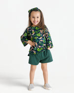A young girl stands smiling in the Jammie Session Bundle of Koi Comfy Cloud Short Set, featuring a matching green floral top, bow headband, green shorts, and light slip-on shoes against a white background.