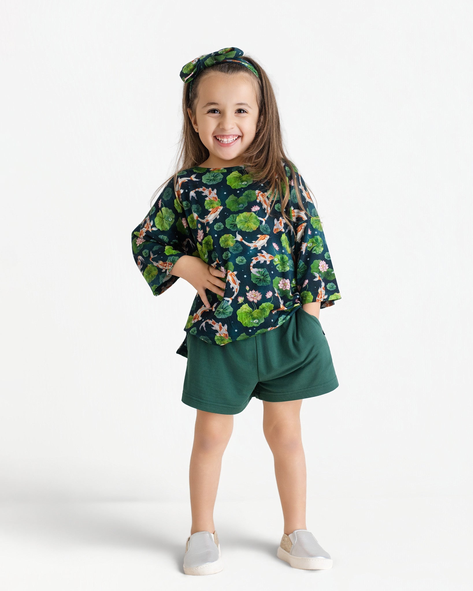 A young girl stands smiling in the Jammie Session Bundle of Koi Comfy Cloud Short Set, featuring a matching green floral top, bow headband, green shorts, and light slip-on shoes against a white background.