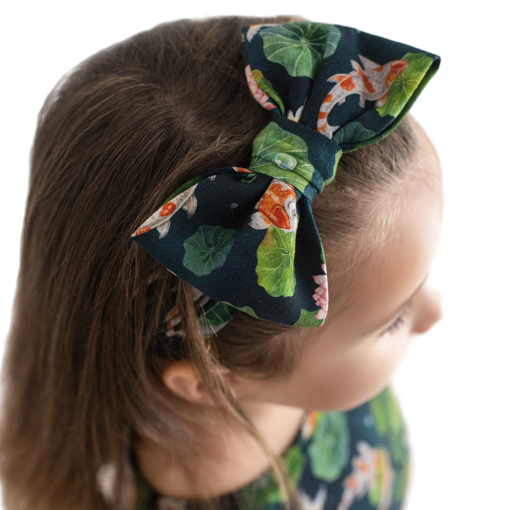 A child with light brown hair wears the Jammie Session Bundle of Koi Bow headband, a large dark blue accessory featuring koi fish and lily pad designs.
