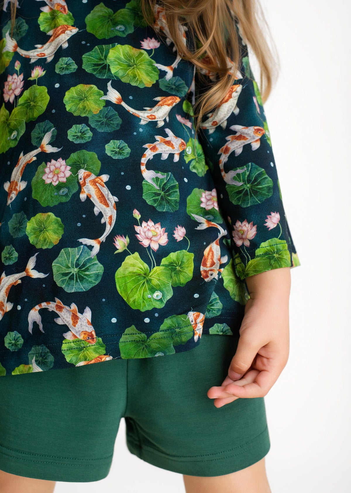 A child wears the Jammie Session Bundle of Koi Comfy Cloud Short Set featuring green shorts and a long-sleeved shirt with a koi fish and lily pad pattern.