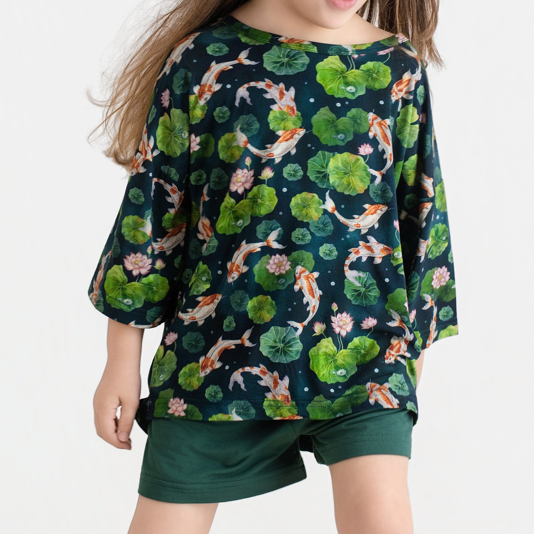 A child models the Jammie Session Bundle of Koi Comfy Cloud Short Set, featuring a dark shirt with a koi fish and lily pad print and matching dark green shorts, against a plain light background.
