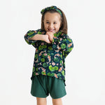 Young child smiles with arms crossed, wearing the Jammie Session Bundle of Koi Comfy Cloud Short Set—featuring a koi fish and lily pad pattern shirt, dark green shorts, and matching headband—against a plain white background.