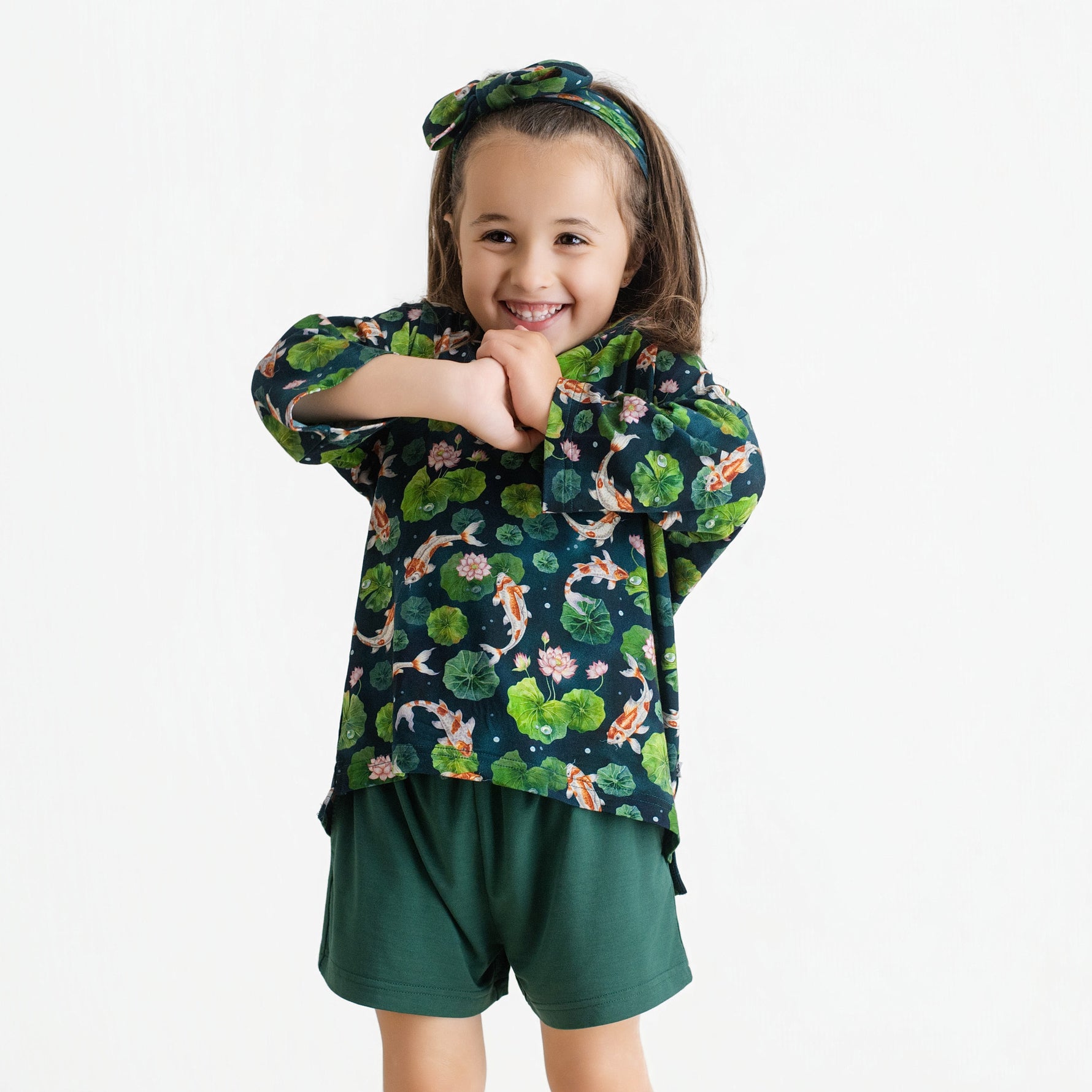 Young child smiles with arms crossed, wearing the Jammie Session Bundle of Koi Comfy Cloud Short Set—featuring a koi fish and lily pad pattern shirt, dark green shorts, and matching headband—against a plain white background.