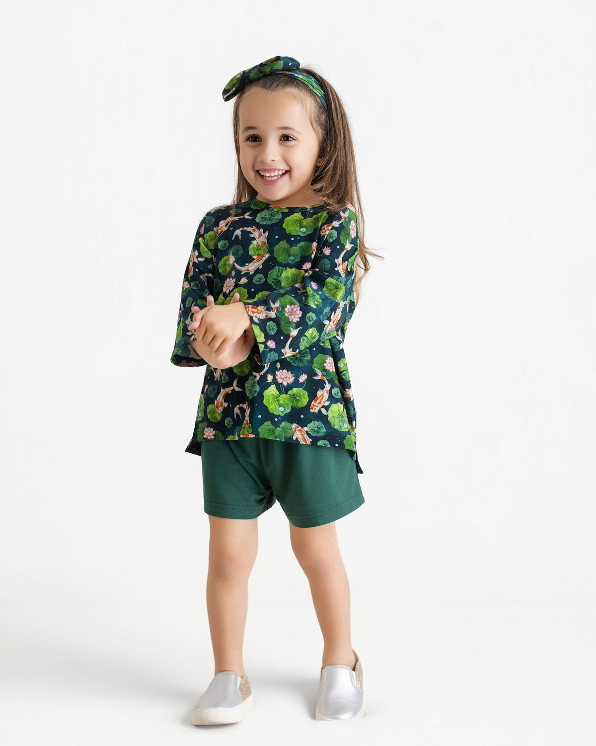 A young girl smiles in the Jammie Session Bundle of Koi Comfy Cloud Short Set—featuring a green floral long-sleeve top, matching shorts, and a floral headband—with silver slip-on shoes, set against a plain white background.