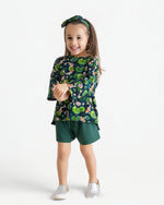 A young girl smiles in the Jammie Session Bundle of Koi Comfy Cloud Short Set—featuring a green floral long-sleeve top, matching shorts, and a floral headband—with silver slip-on shoes, set against a plain white background.